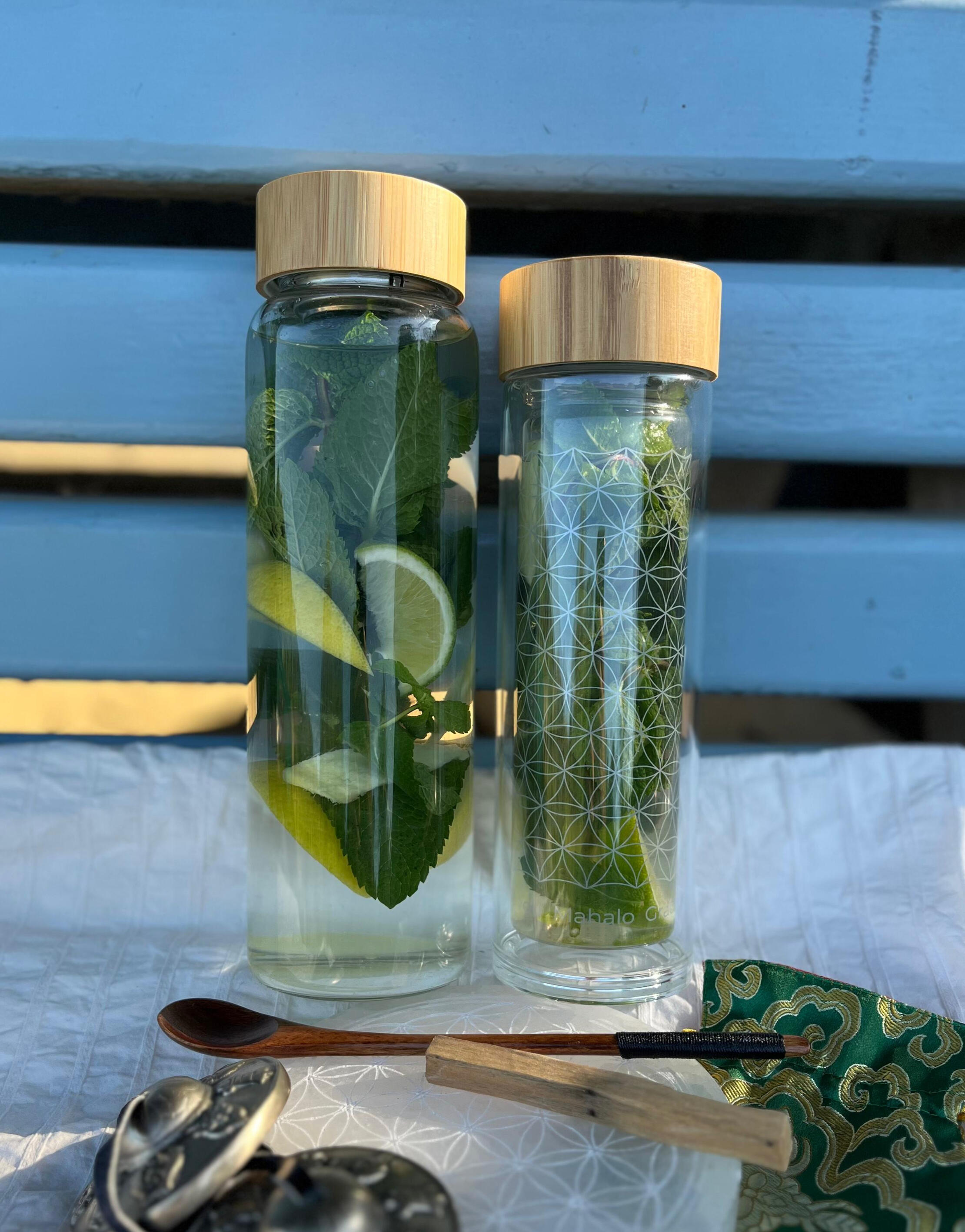 Two AMAZI glass tea bottles with Flower of Life engraving on blue bench, filled with fresh herbs and lemon water, surrounded by ritual tools including wooden spoon and sacred geometry cards