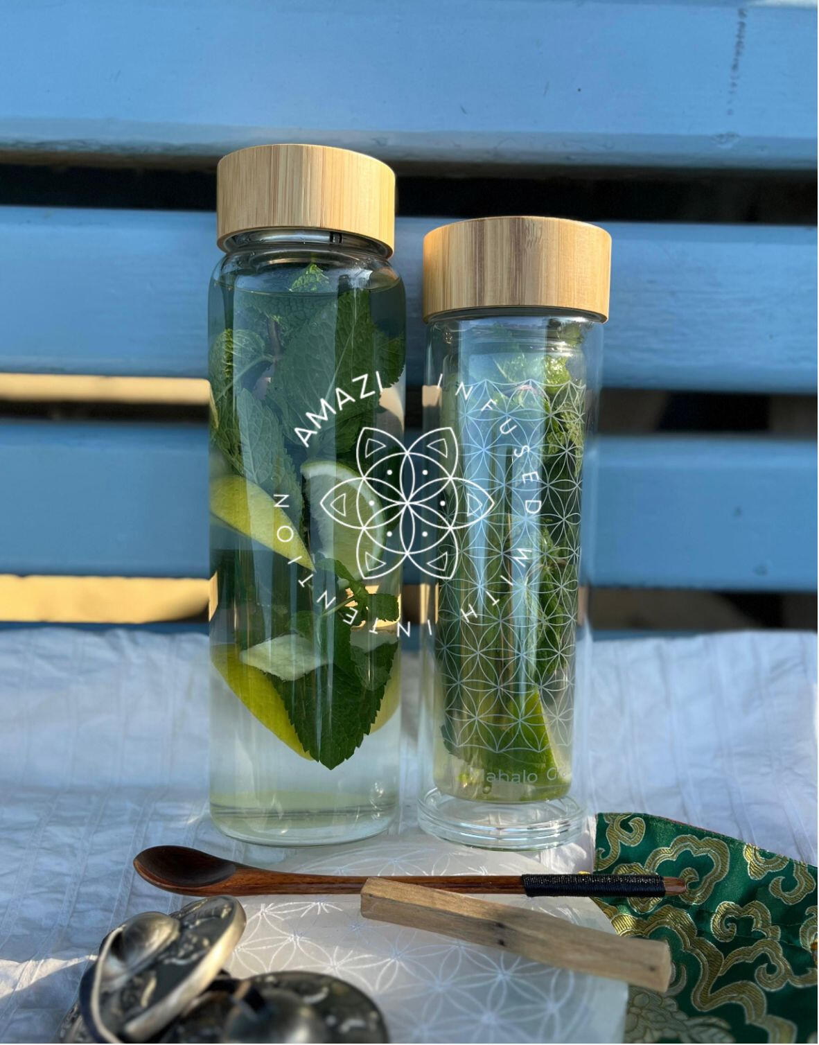 AMAZI Sacred Tea Bottles with Flower of Life Design Two AMAZI glass tea bottles with Flower of Life engraving on blue bench, filled with fresh herbs and lemon water, surrounded by ritual tools including wooden spoon and sacred geometry cards